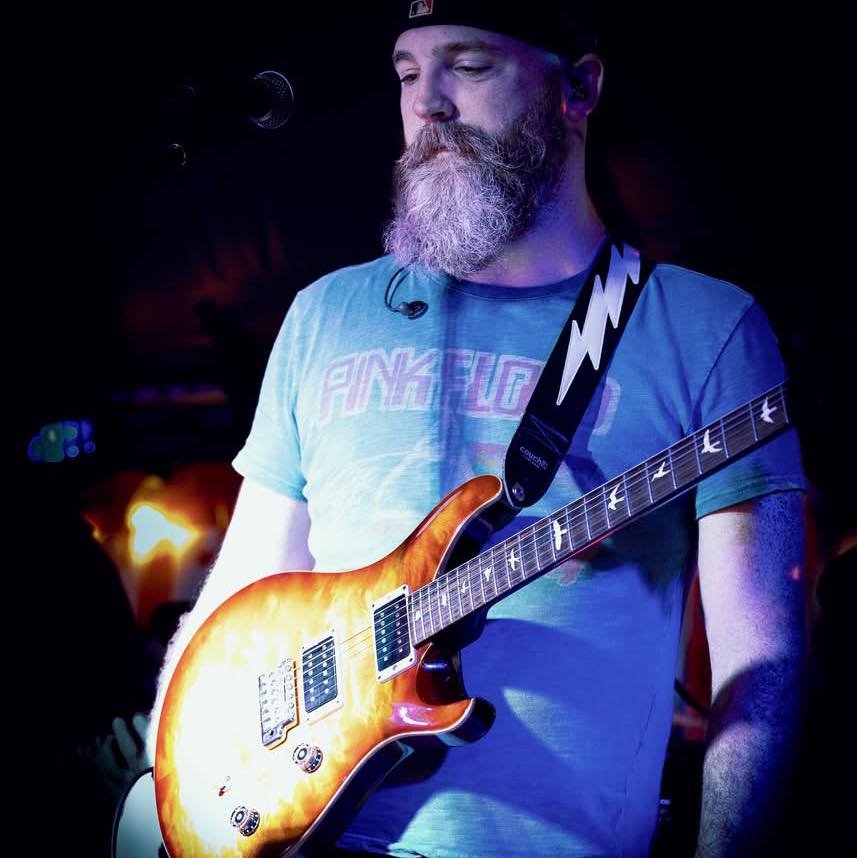 Photo of Dan in full color with a black background. Dan is wearing a blue t-shirt with a design featuring the band Pink Floyd. He is also wearing an electric guitar. His guitar strap is black with a white lightning bolt on it, the guitar is a PRS CE24 in Livingston Lemondrop.